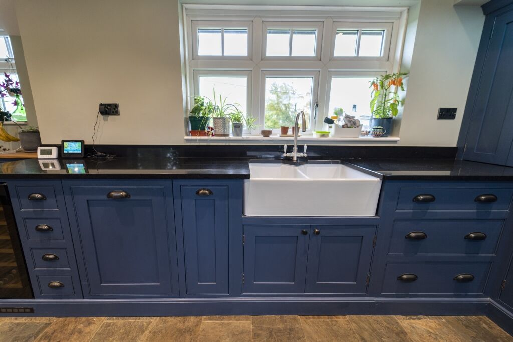 Front view of a blue kitchen sink run with Absolute Black Granite worktops, showing a white farmhouse sink, tap and window plants above the worktop.