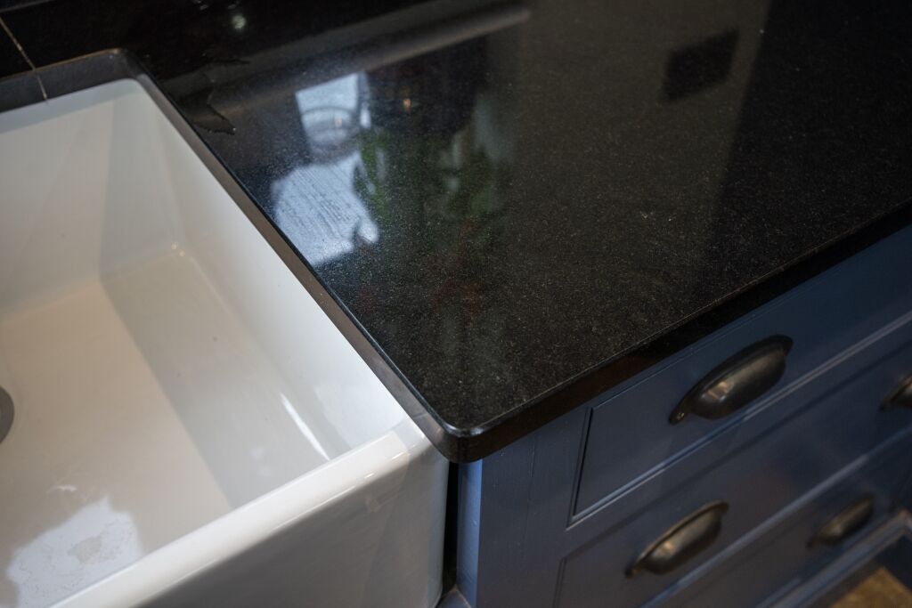 Close up of an Absolute Black Granite worktop corner with a polished finish, showing the edge detail above blue cabinetry next to a white sink.