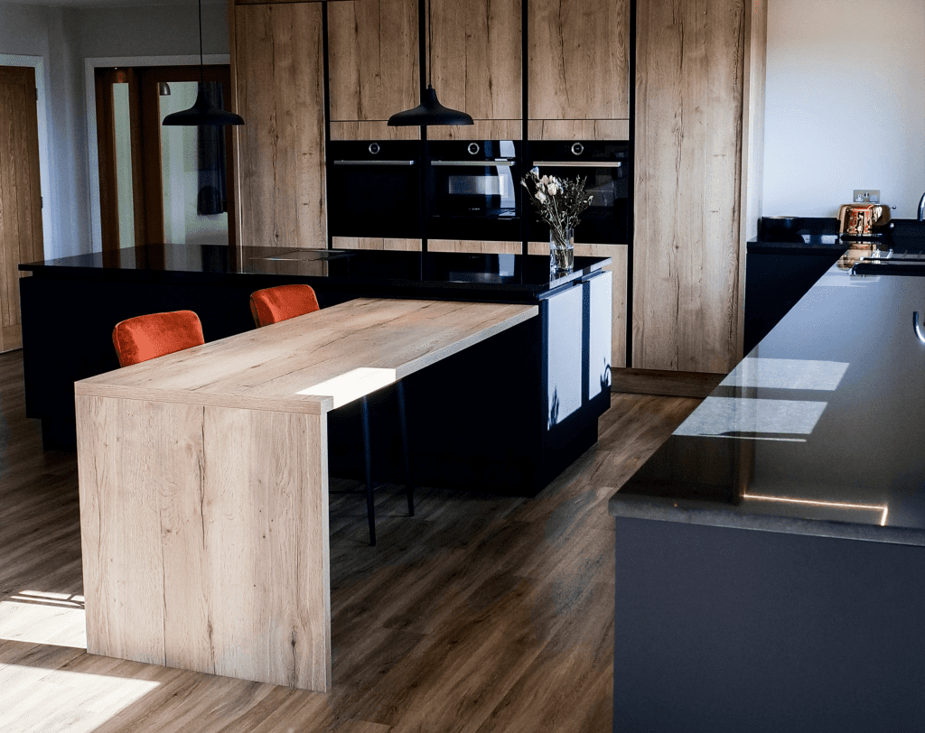 Wide kitchen view showing an Absolute Black Granite island worktop with a long breakfast bar and timber cabinetry, with pendant lights and seating.