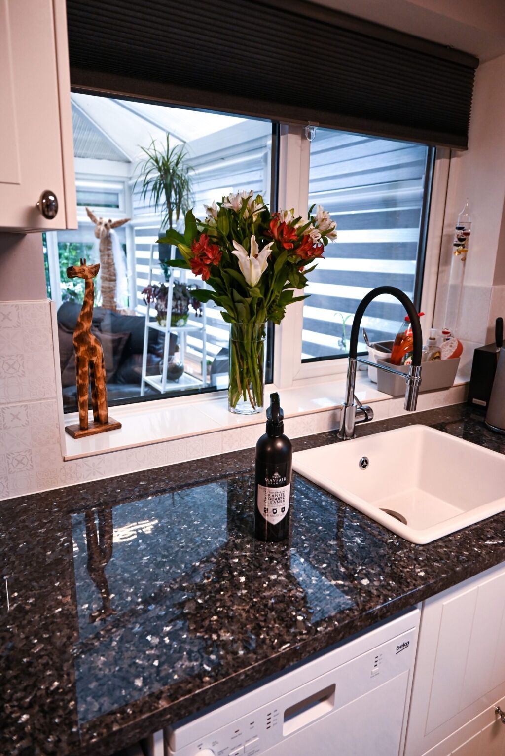 Corner sink view with Blue Pearl Granite worktops, showing the polished speckled Granite surface, black tap and flowers beside a window.