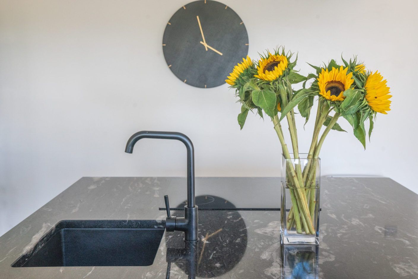 Close up of a sink area in Cosmic Black Granite with a black tap, showing the polished reflective surface and sunflowers in a vase on the worktop.
