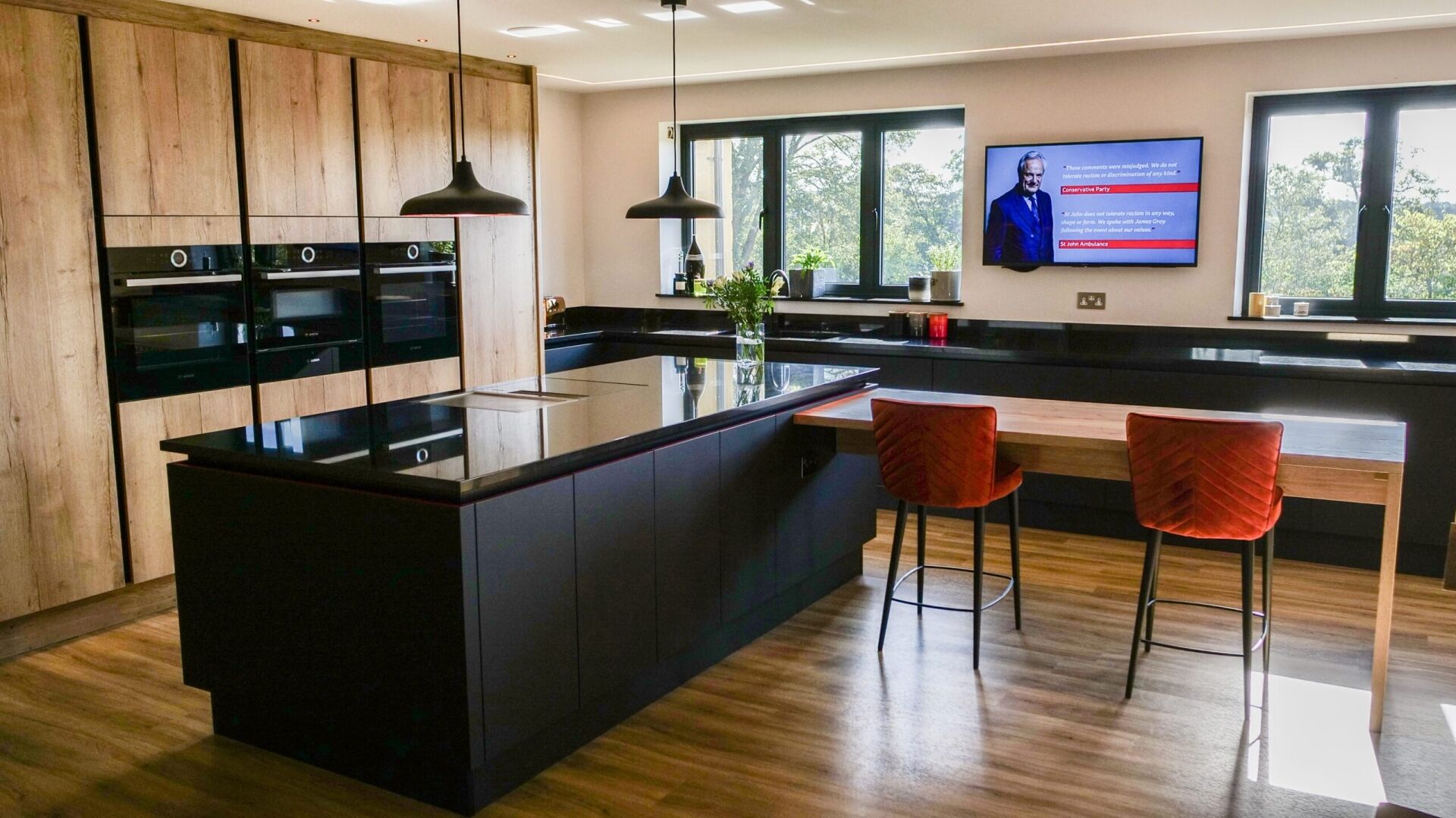 Wide view of a modern kitchen with an Absolute Black Granite island worktop and breakfast bar, showing bar stools, pendant lights and large windows.
