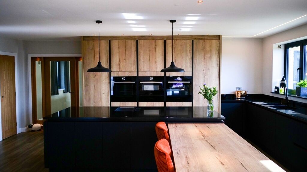 Kitchen view showing an Absolute Black Granite island worktop with breakfast bar and seating, with timber wall units and built in ovens in the background.